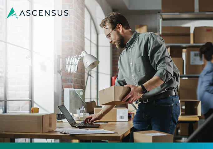 Ascensus logo above a small business owner packing boxes at a desk in a warehouse-style shop, working on a laptop.
