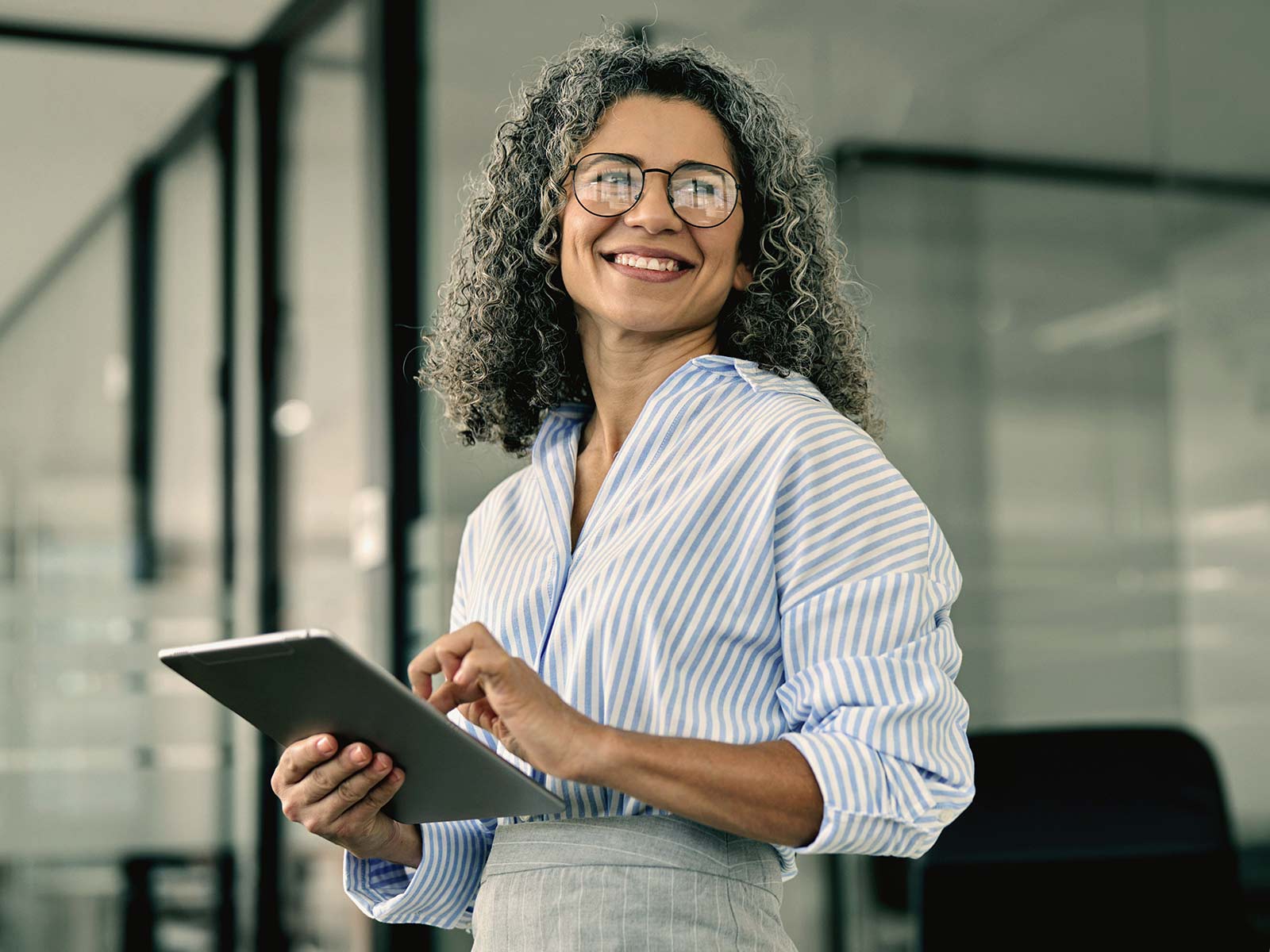 Smiling businesswoman with curly gray hair using a tablet in a glass-walled office