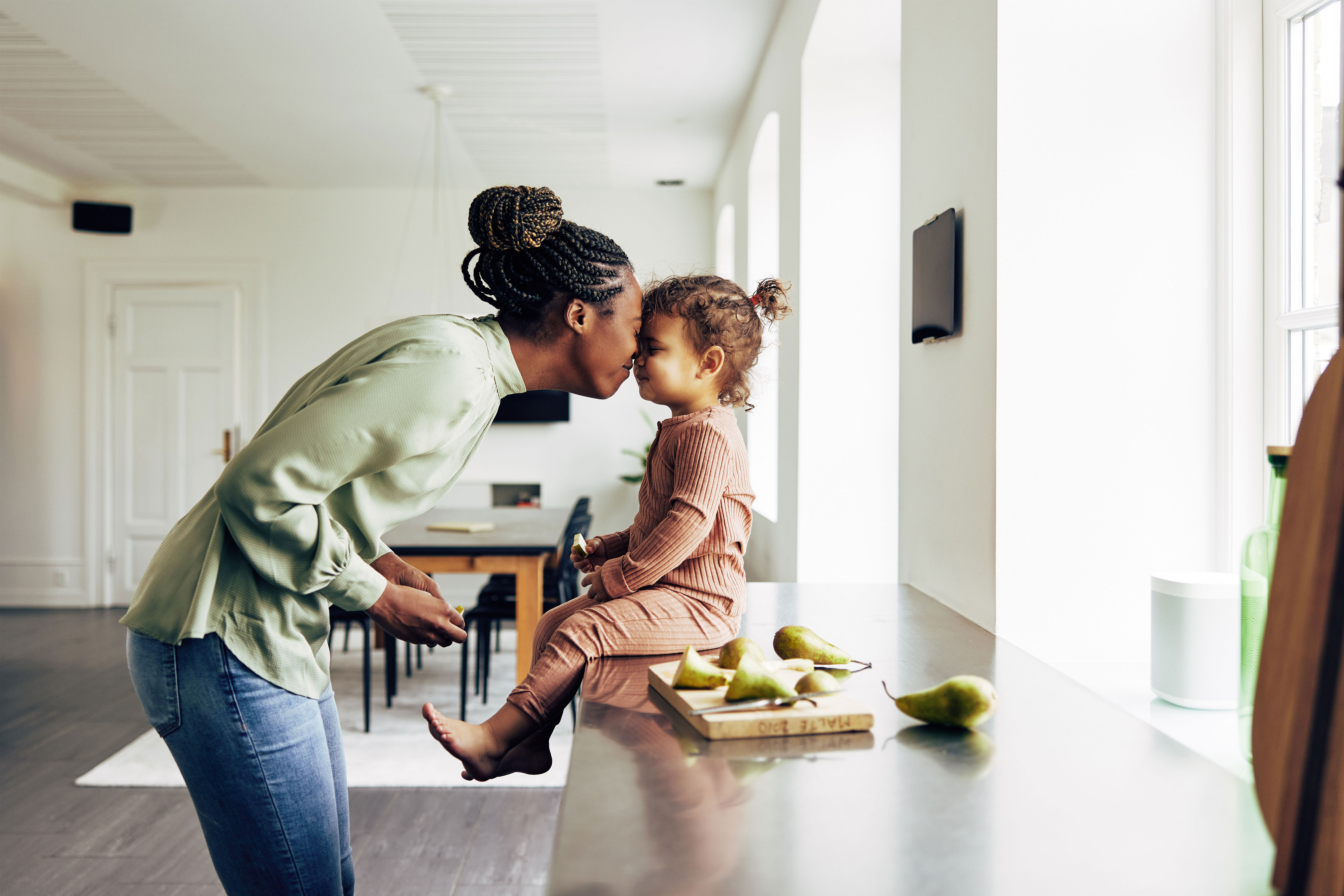 A mother and young child touching noses while sitting on a kitchen counter with sliced pears