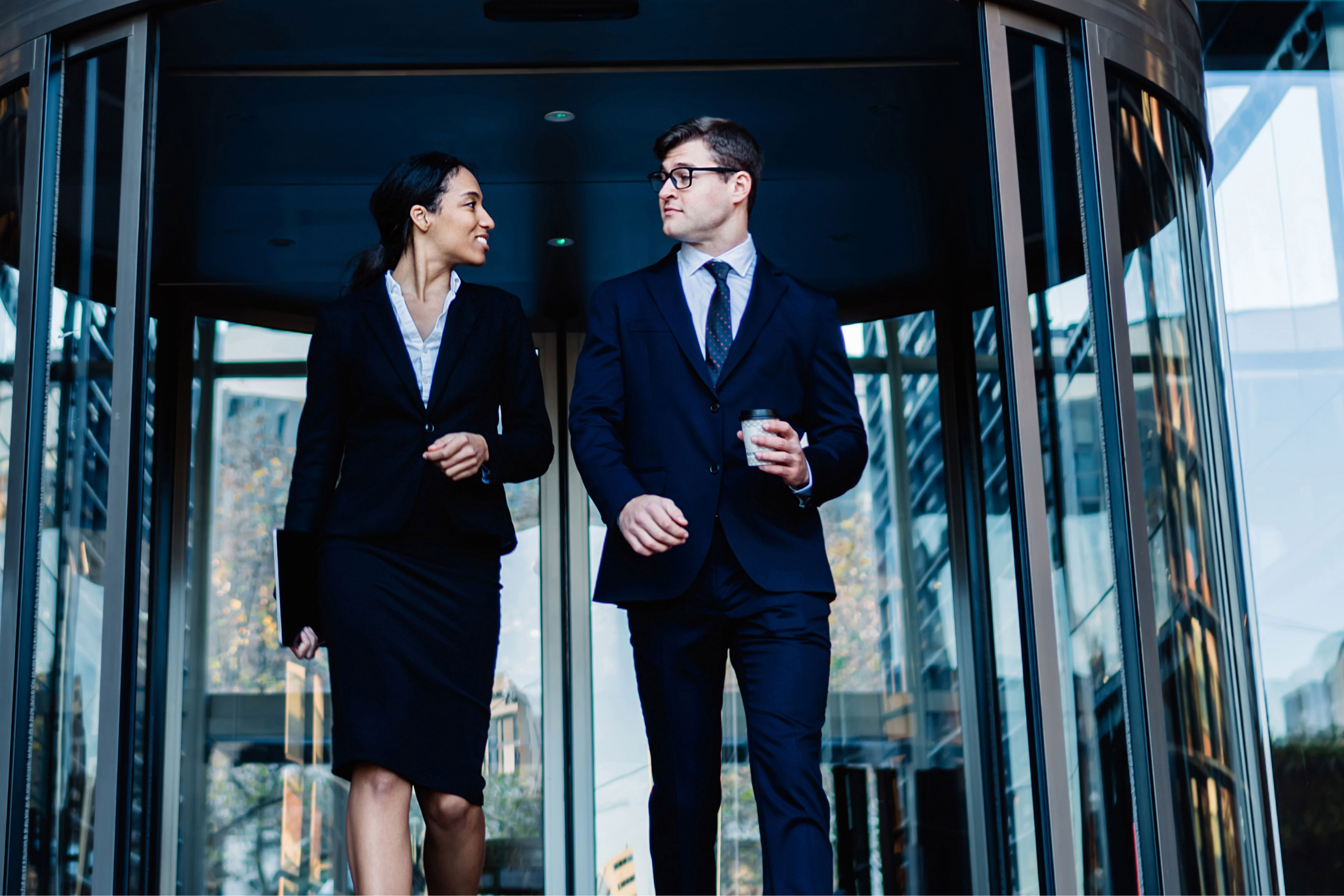 Two business professionals in suits conversing while walking through an office building's revolving door entrance